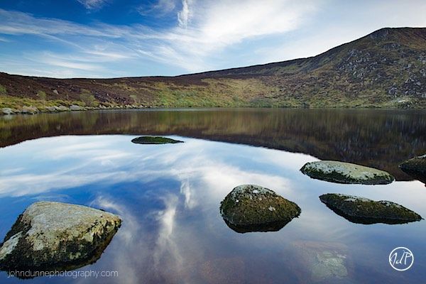 As the sun sets behind the mountains of Wicklow Lower Lough Bray falls into a stillness with the clouds above reflecting off the mirror like lake, and just the cliffs and old stones standing guard for the coming night.