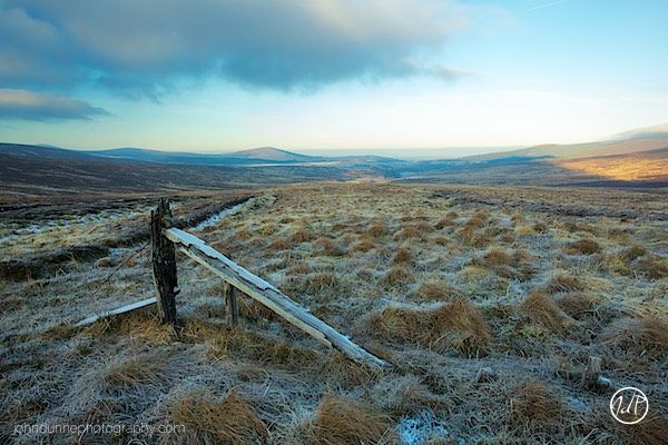 A roughly built fence lies in a frost covered field as the sun rises to shine upon the mountainous Wicklow landscape of Ireland.