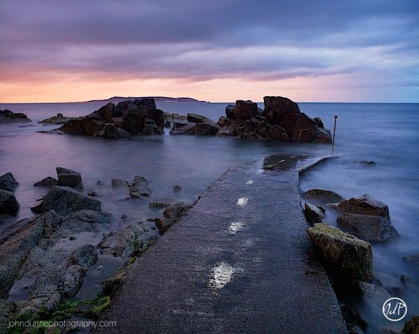 The last light of the day throws its soft amber light through the heavy clouds above Howth as the viewer stands upon the slip in Sandycove.
