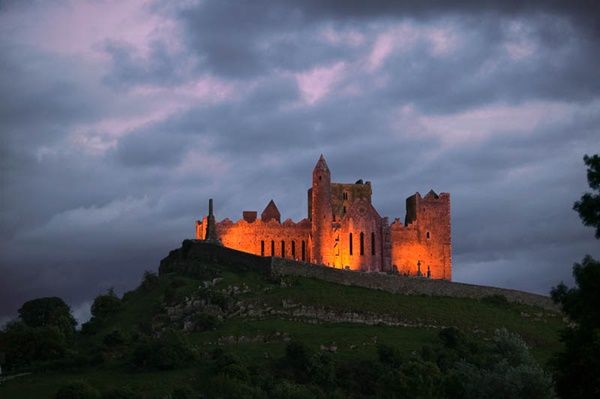 Midlands of Ireland: Tipperary: Cashel. This image is a photograph of The Rock of Cashel after sunset. There is still a little light left in the sky.  Cashel, also known as Cashel of the Kings, is dominated by the huge and iconic castle known as The Rock of Cashel (Carraig Padraig or St Patrick's Rock, as it was formally known). It is reputedly the site of the conversion of Aenghus the King of Munster by St. Patrick in the 5th century AD.  Long before the Norman invasion of Ireland, The Rock of Cashel was the seat of the High Kings of Munster.  Most of the buildings on the current site date from the 12th and 13th centuries when the rock was gifted to the Church. It is one of the most remarkable examples of medieval architecture to be found anywhere in Europe.