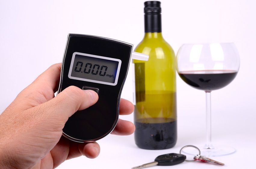 Checking someones blood alcohol level with a small breathalyser. A red wine bottle, wine glass and car keys are in the background.