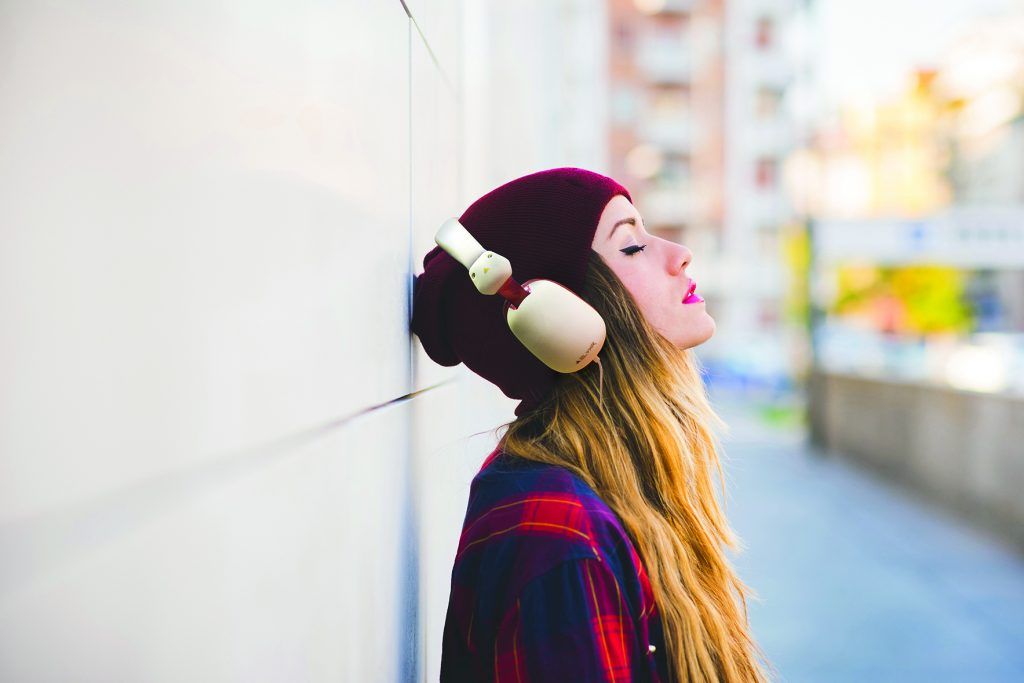Half length profile portrait of young handsome caucasian blonde hair woman leaning against a wall, listening music with headphones, eyes closed - serene, enjoying, music concept